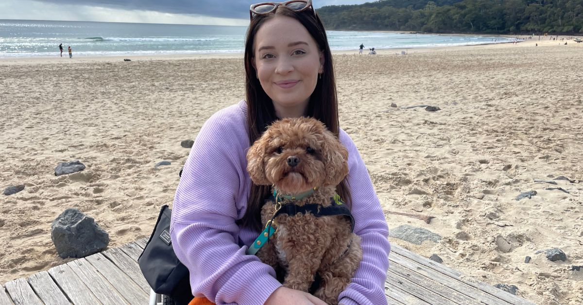 A young woman with fair skin, long, dark hair and sunglasses perched on her head, wearing a purple jumper and holding a small service dog on the beach. She is smiling for the camera.
