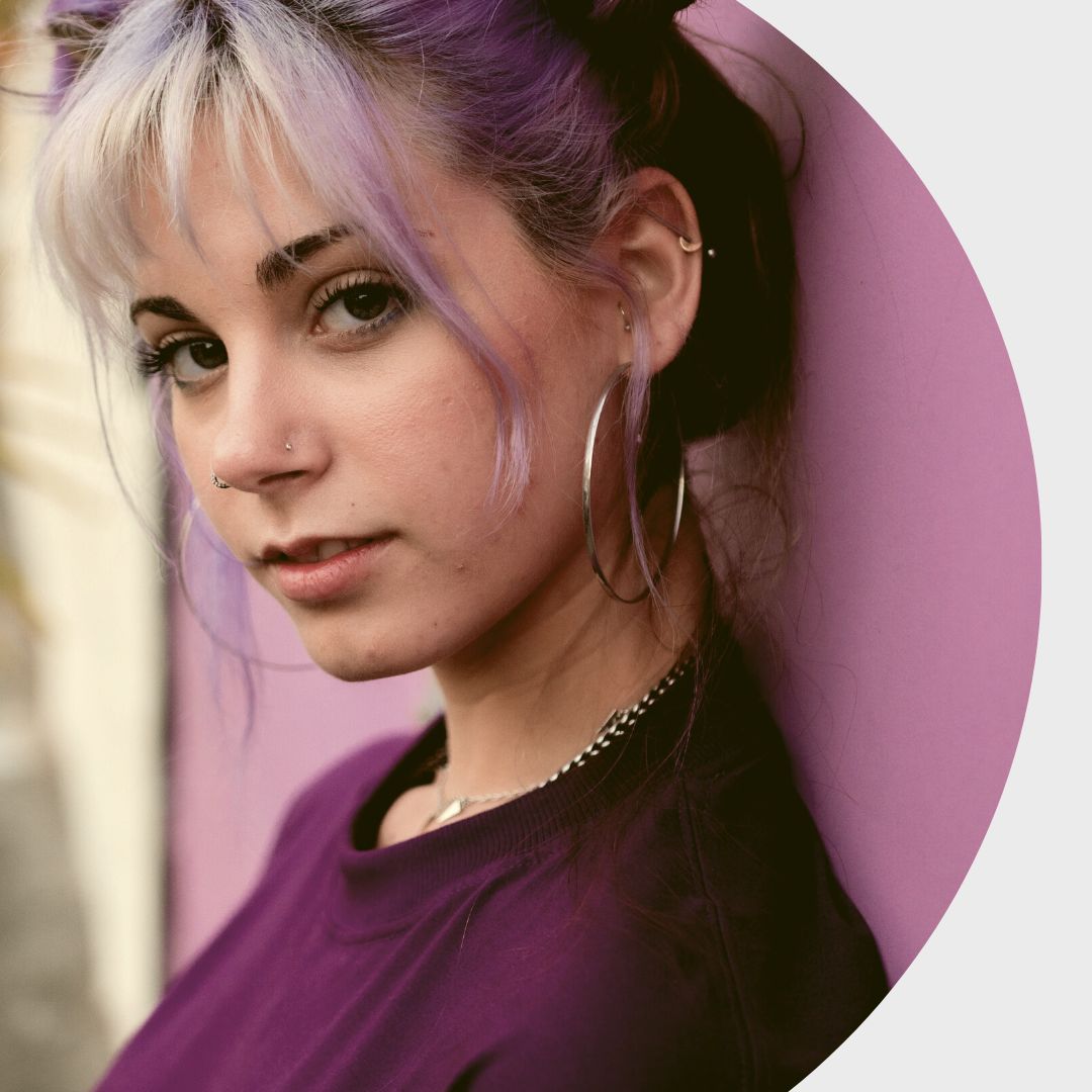 A young woman with pink and blond hair, heavy make-up and hoop earrings wearing a purple jumper and leaning on a pink wall, looking at the camera with a serious expression.