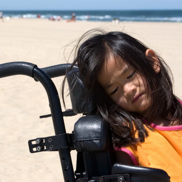 A little girl with shoulder length black hair, fast asleep in a motorised wheelchair. The beach can be seen behind her.