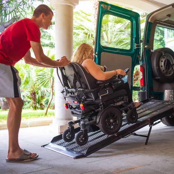 A man pushing a young woman in a motorised wheelchair up a ramp and into the back of a green van. The man wears a red t-shirt and grey shorts, the woman has long blond hair. They are in an undercover parking area or loading bay with trees in the background.