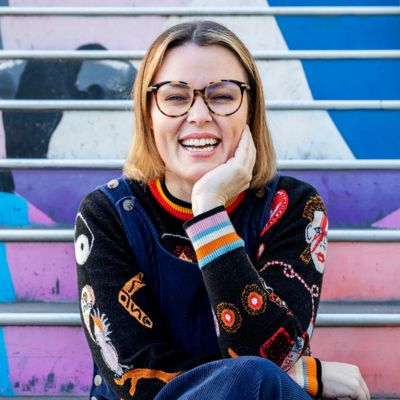 A photograph of a young woman with short, dyed red hair, fair skin, glasses and a big smile, wearing a brightly coloured jumper with jeans, sitting at the bottom of a brightly coloured set of steps with her chin in her hand and elbow resting on her knee.