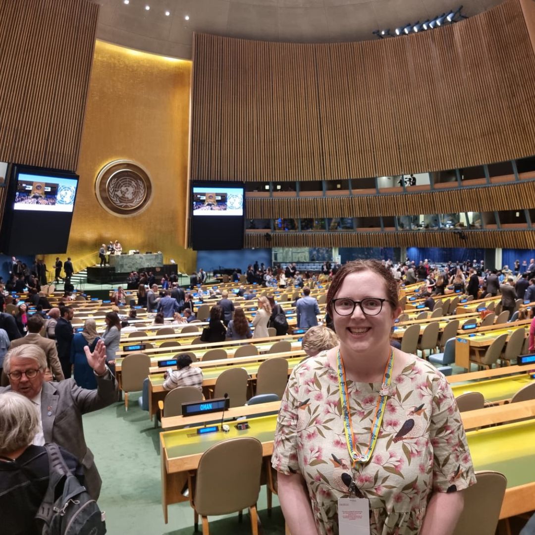 A young, smiling woman with fair skin, brown hair tied back and glasses, wearing a floral dress, standing in a large auditorium filled with rows of chairs at long yellow benches. The UN logo is lit up on the far wall.