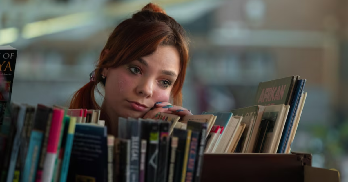 A photograph of Quinni from Heartbreak High. She is standing behind a bookshelf, resting her hands and head on a line of books. She has auburn hair and looks thoughtful.