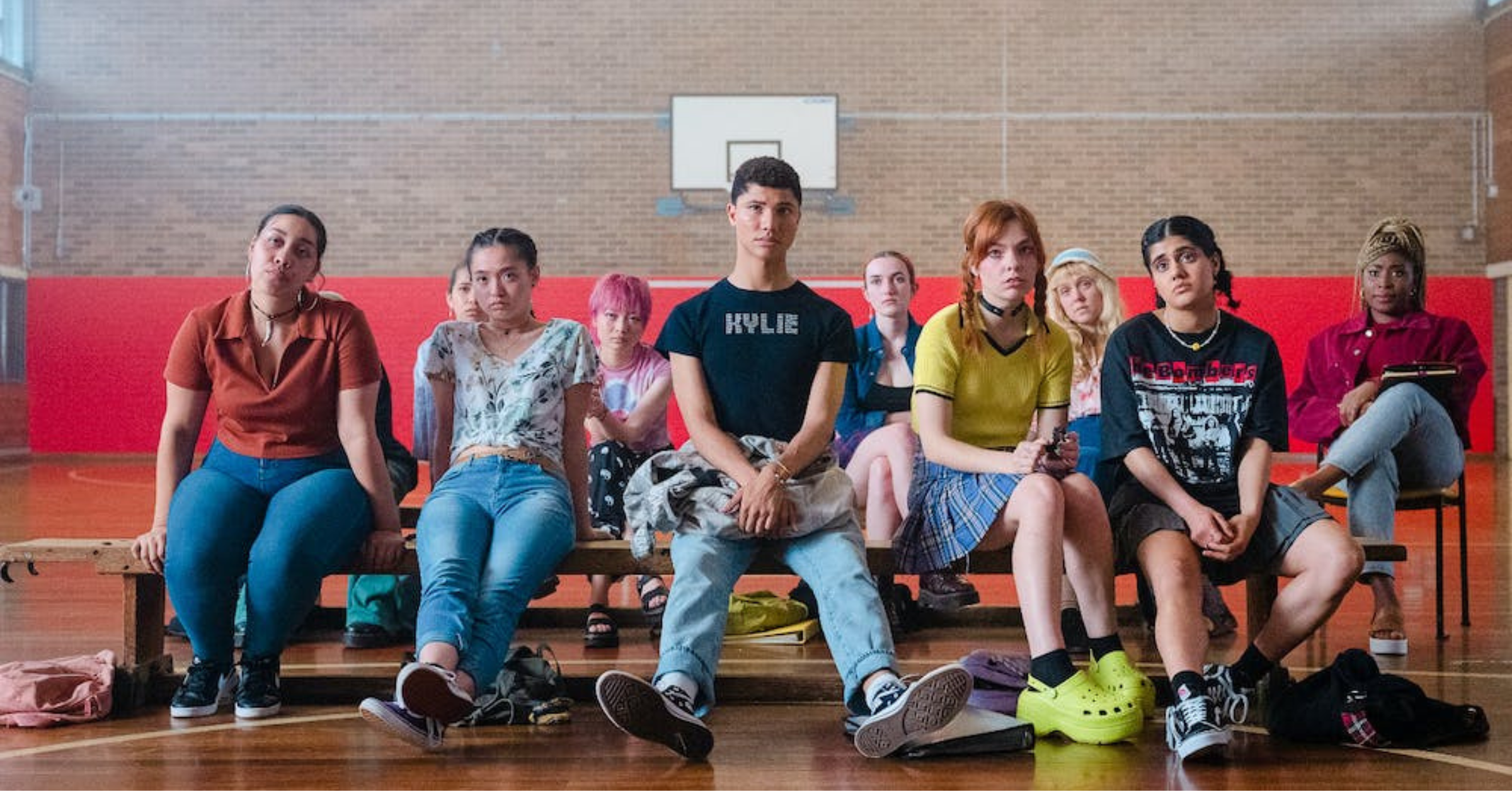 A still image from the Netflix show Heartbreak High shows a group of about a dozen teenagers sitting in their school's basketball court. The character in the middle front is wearing a black T-shirt with the word 'Kylie' emblazoned on the front, as well as light blue denim jeans. To their right is a young woman wearing a yellow top and green crocs. To her right is another woman wearing a black shirt and sneakers.