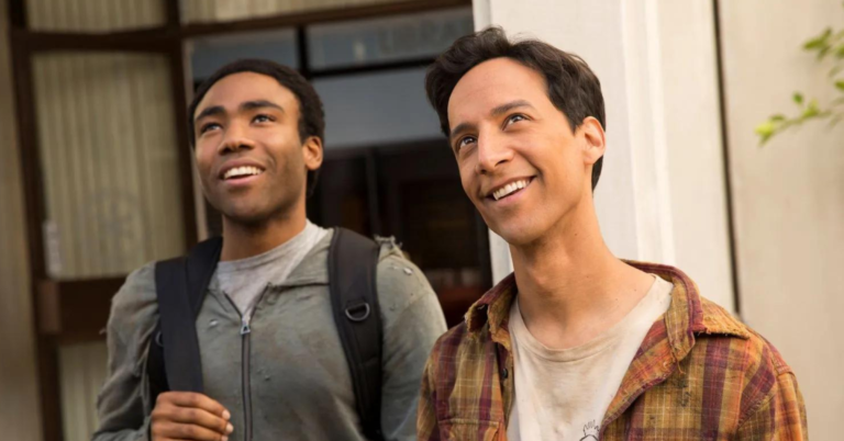 A still from the TV series Community shows Abed and his friend Troy looking up at something while smiling. Abed, on the right, has dark brown hair and is wearing an orange and red shirt. Troy, to his left, is dark-skinned, has close-cropped black hair and is wearing a grey hoodie with the zipper done up. Behind them is the entrance to a building.
