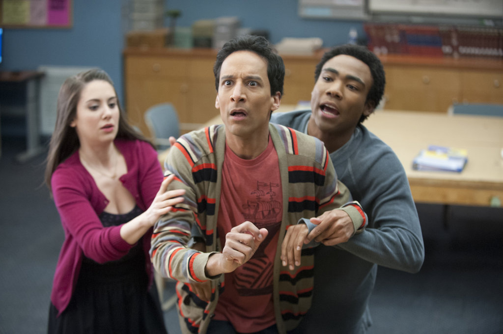 A photograph shows Abed, a man with brown skin and black hair wearing a red shirt and colourful cardigan, being restrained by his two friends. To his left is Annie, a brown-haired woman wearing a pink cardigan, and to his right is Troy, a dark-skinned man with black hair and a grey-blue sweater. 