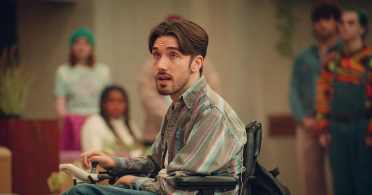 A still image from the show Sex Education shows the character Isaac. He has wavy brown hair, a moustache and stubble, and is wearing a pink, blue and grey collared shirt. Isaac is in the middle of a classroom in front of a crowd of blurred students and is using an electric wheelchair.