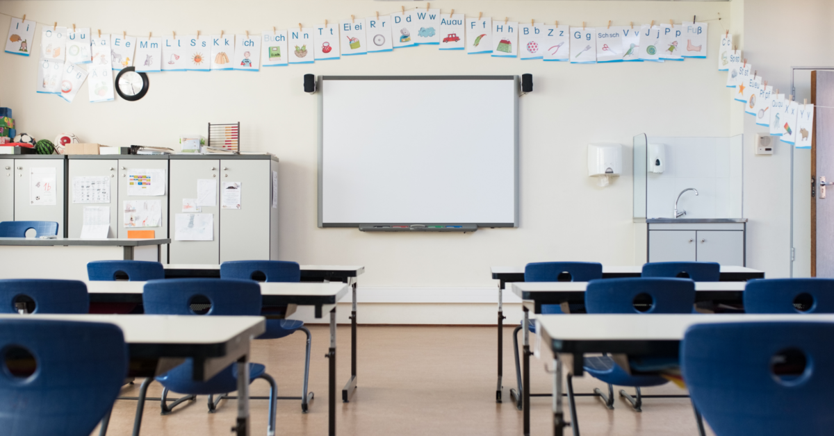 A photograph shows an empty classroom. A series of desks and blue chairs are facing a white board at the front of the room. Small posters showing letters of the alphabet hang on a string above the board.