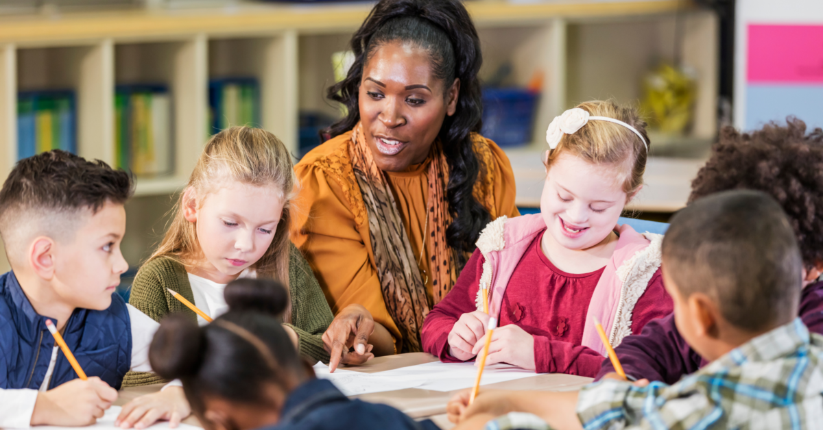 A photograph shows a teacher with long brown hair in an orange top. She is surrounded by a group of students in colourful clothing who are writing on sheets of paper using pencils. They are in a classroom.