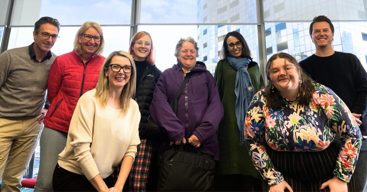 8 smiling people of varying age and gender, all in smart casual clothes, posing together in front of an office window. The two women in front are hunched down a little so the people in the back can be seen. The photo is taken from low down, looking up at the group.