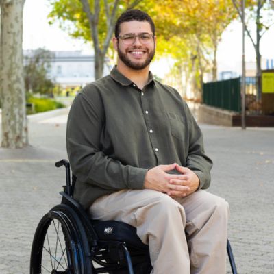 A photo of a young man with olive skin, glasses, a short beard and a big smile, sitting in a manual wheelchair with his hands clasped in his lap. He is wearing khakis and is in an outdoor area with concrete paths and trees.