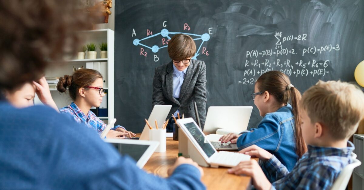 A photograph shows a group of children in a classroom in front of a blackboard covered in equations. The five children are using laptops and sitting around a central table. The back of the a teacher can be seen in the foreground of the image supervising the class.