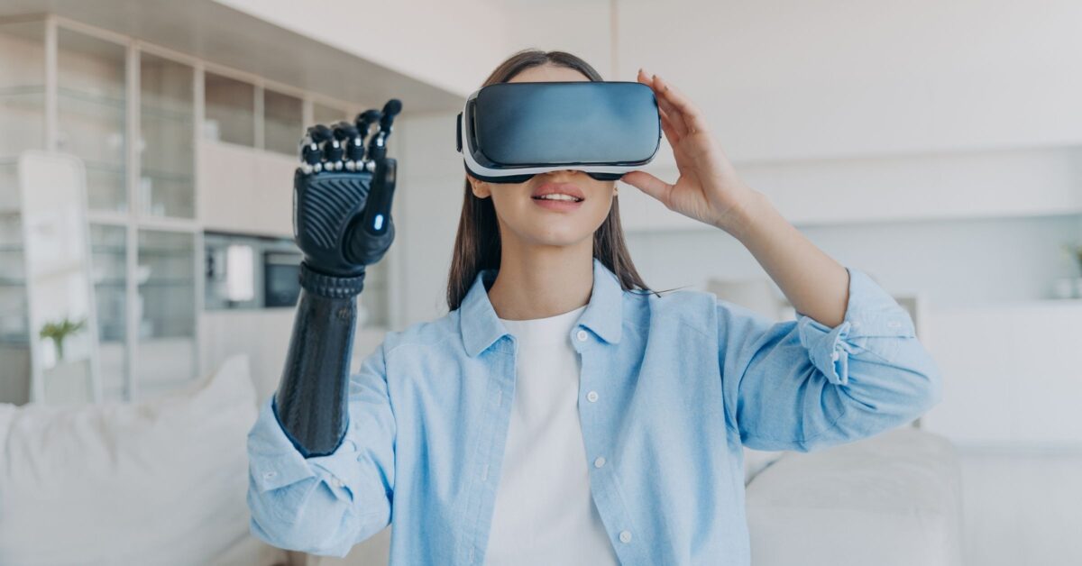 A photograph shows a woman with long brown hair in a white t-shirt under an open light blue button-up shirt. The woman has a black prosthetic right arm and is wearing a virtual reality headset.