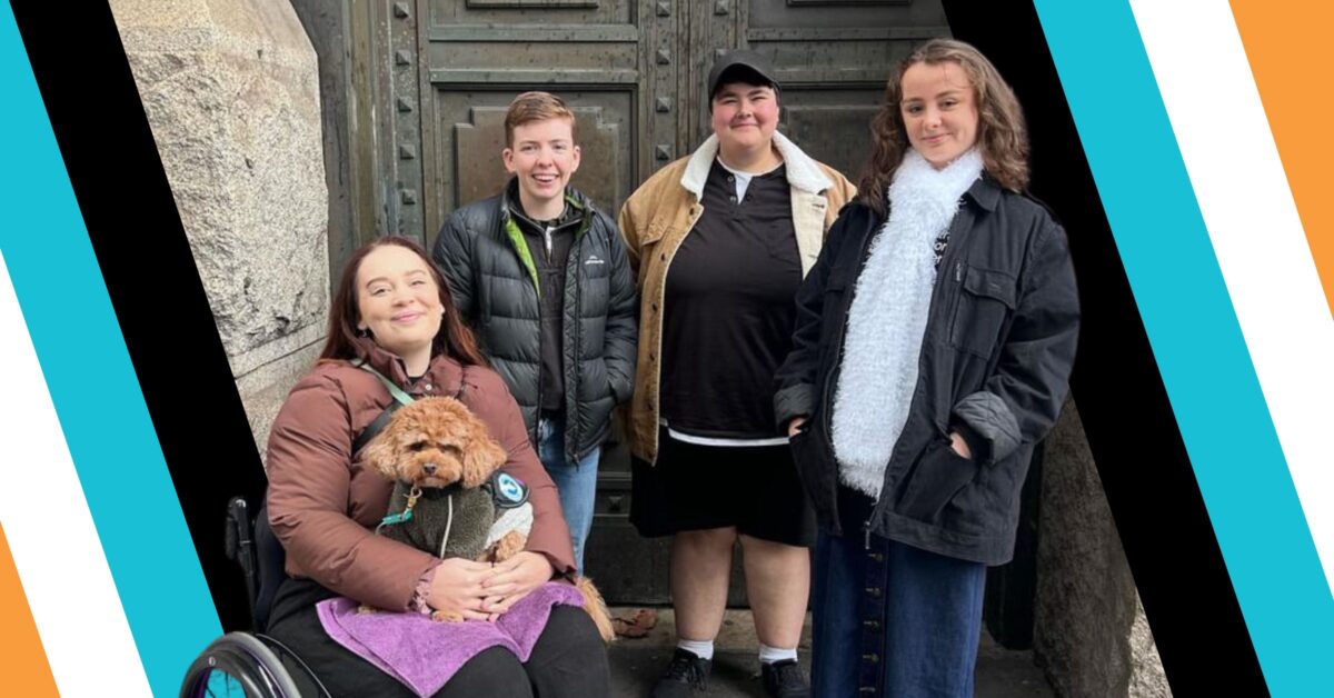 A photograph shows a group of four young people in front of a pair of iron metal doors. They are all smiling at the camera. The young person on the left is using a wheelchair and has a small brown dog on her lap. The image is framed by a thick tilted border coloured orange, white, light blue, and black.
