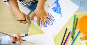 A photograph shows a young child using a paintbrush and blue paint to draw an outline around their left hand.