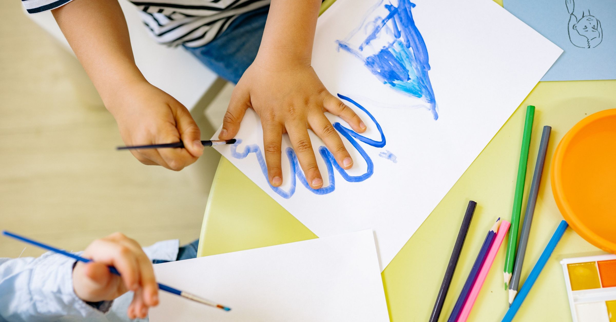 A photograph shows a young child using a paintbrush and blue paint to draw an outline around their left hand.