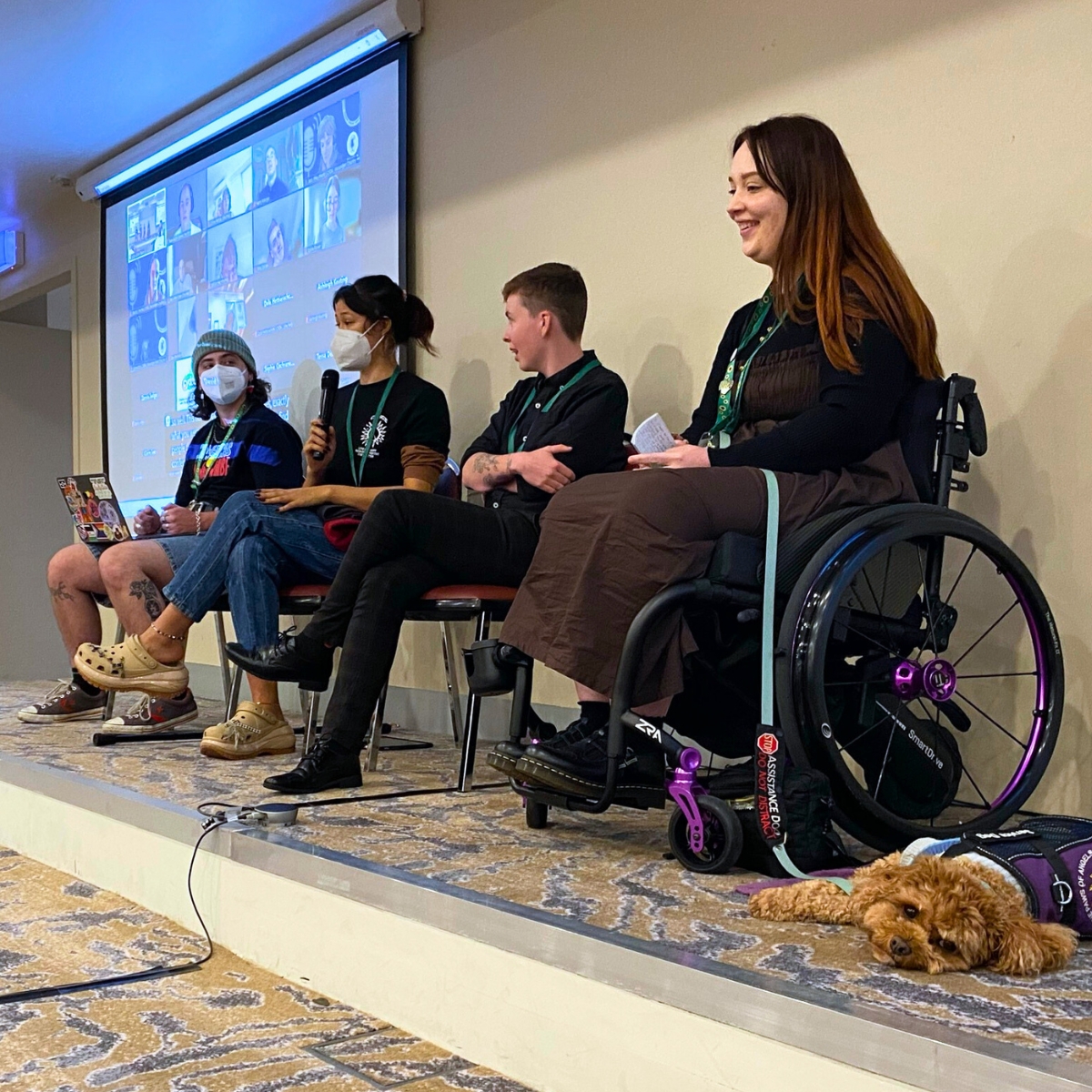 A panel of four young people speaking from a stage. The woman on the right is using a manual wheelchair and has a service dog lying next to her. The two people on the left are wearing face masks. Behind them is a projector screen with lots of people in a Zoom meeting.