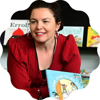 A smiling woman with dark hair wearing a red cardigan with puffy sleeves and holding a children's picture book titled "Come over to my house". There is a shelf full of children's books behind her.