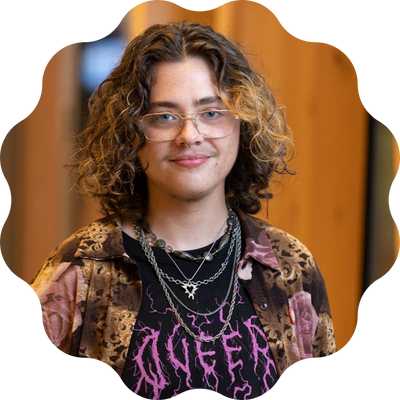 A young person with shoulder length, dark, curly hair with highlights wearing a lot of chain necklaces, and a t-shirt with "Queer" printed on it in pink letters shaped like lightening.