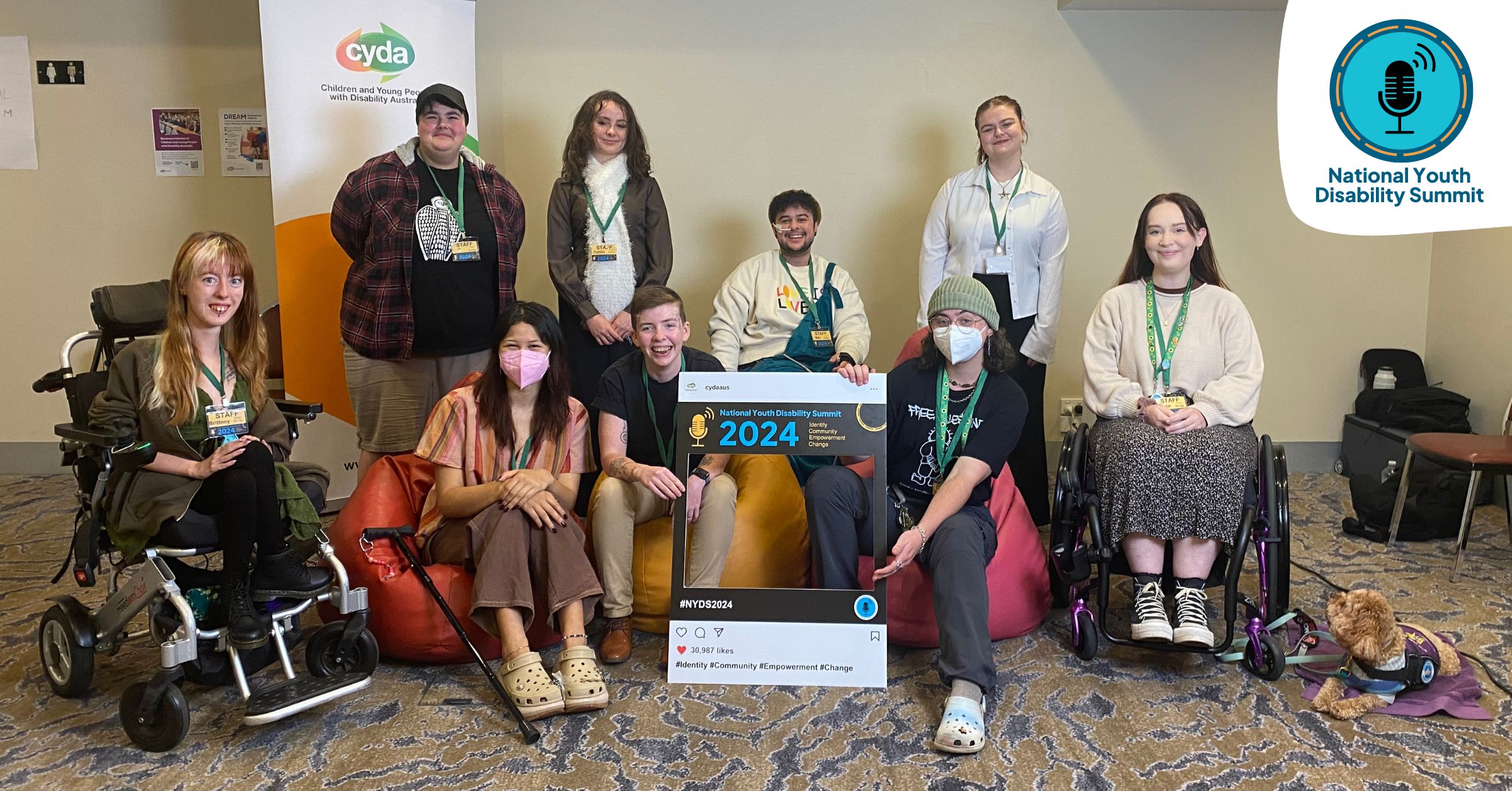 A photo of some of CYDA’s Youth Council in a well-lit room in front of a green, orange and white banner featuring the CYDA logo. Three people at the front of the group, including Ezra on the right, are sitting on colourful beanbags. Ezra and another young person are holding a cardboard cutout made to look like an Instagram post with the text “National Youth Disability Summit 2024.” The Summit's logo sits top right.