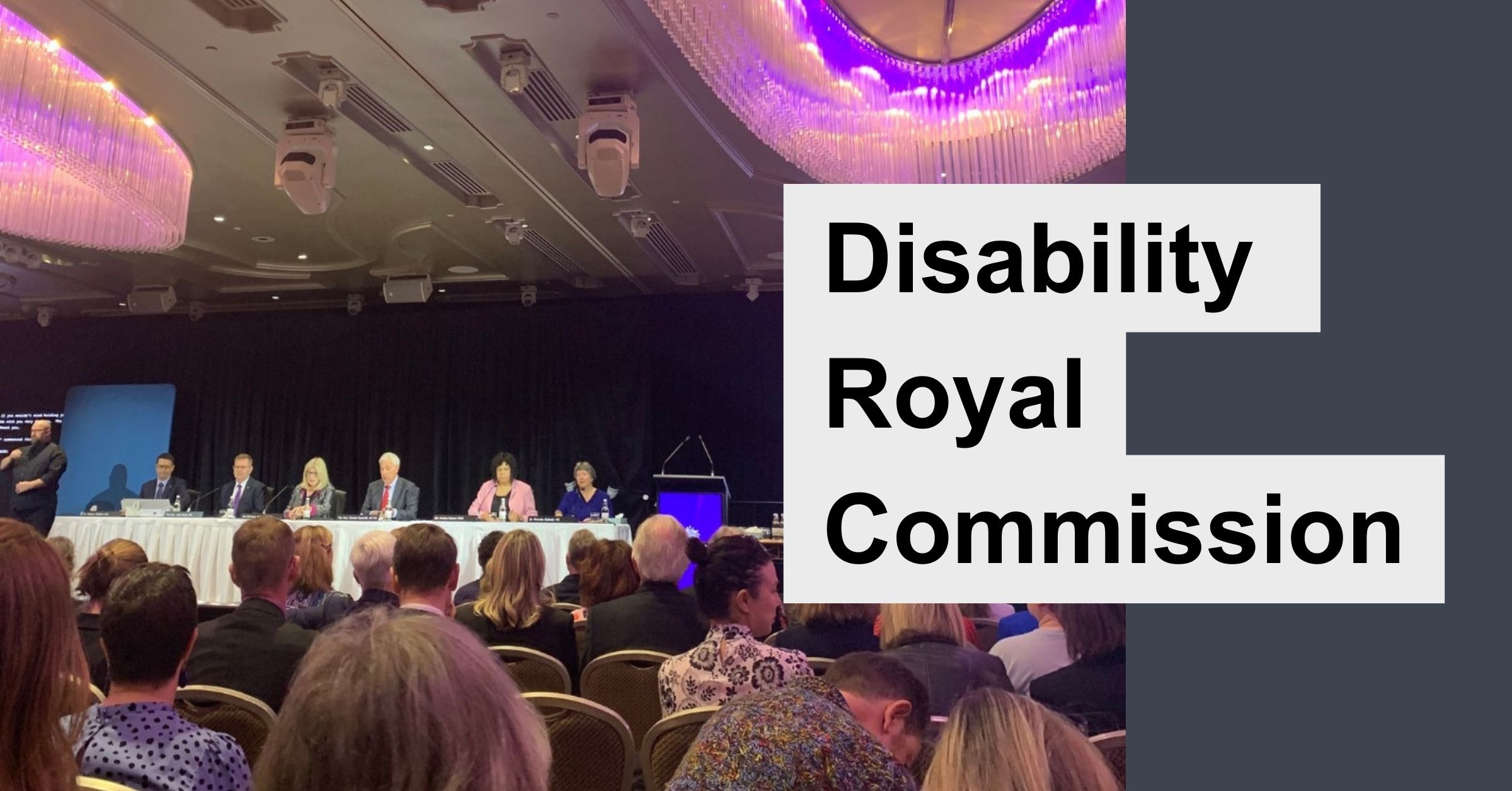 A photo of a panel of six people in suits and dresses sitting at a long desk in a conference room. An audience sits in front of them. Text to the right reads "Disability Royal Commission".