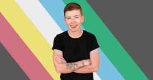 A photo of a smiling young person with short light brown hair wearing a black t-shirt. They have their arms crossed in front of them. A tattoo of a flower sits on their right arm. Behind them is the red, yellow, white, blue, and green Disability Pride flag.