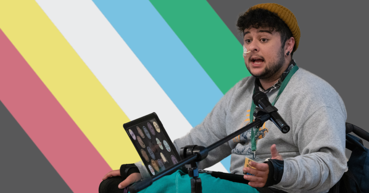 A photo of a young man with curly dark hair using a wheelchair. He is wearing a grey sweater and a brown beanie. The Disability Pride flag takes up the background behind him.