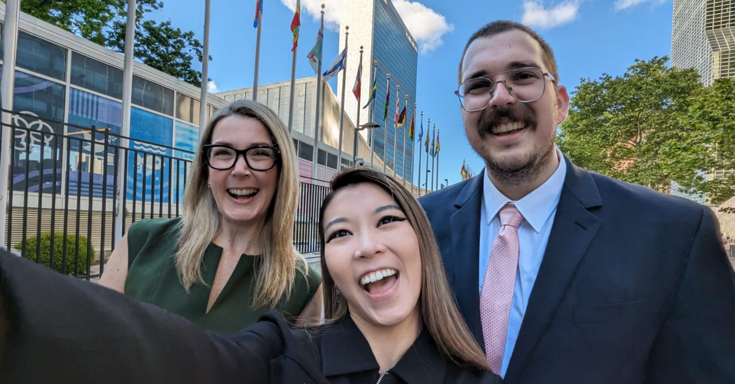 A photo shows three smiling people in front of the United Nations headquarters building. The woman on the left has long blonde hair and is wearing black-framed glasses and a green dress. The person in the middle has long brown-blonde hair and a black jacket. The man on the right has short-cropped brown hair and is wearing a navy suit, a pink tie, and silver-framed glasses.