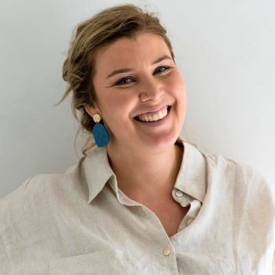A profile photo of a young fair skinned woman with blond hair, a wide smile, and large blue earrings wearing a cream, linen open-collared shirt.