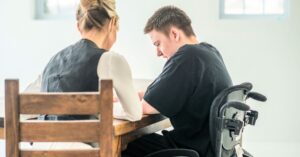 A photo of a teenage boy with short dark blonde hair in a black t-shirt using a wheelchair next to a blonde woman in white and black clothing. The pair are sitting together at a wooden desk.