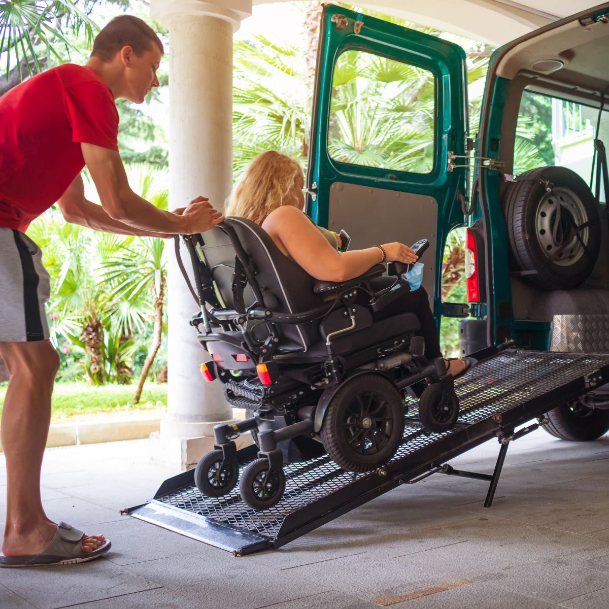 A young woman with curly blond hair using a power wheelchair. A young man in a red shirt and shorts is pushing the wheelchair up a ramp in the back of a green van.