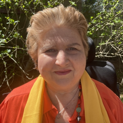 A profile photo of an older woman with short, styled, grey and red hair, wearing an orange shirt with a yellow summer scarf and nice make-up. She is outside on a sunny day with greenery behind her.