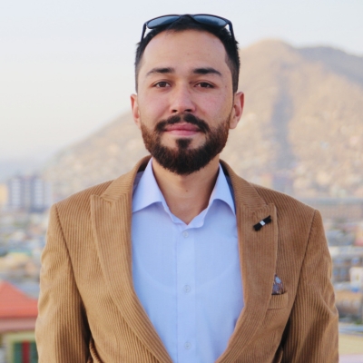 A young, smiling man with olive skin, short dark hair and a neatly trimmed beard wearing a light brown corduroy jacket over a powder blue collared shirt. He is looking directly at the camera, and behind him, out of focus, are pyramids and houses.