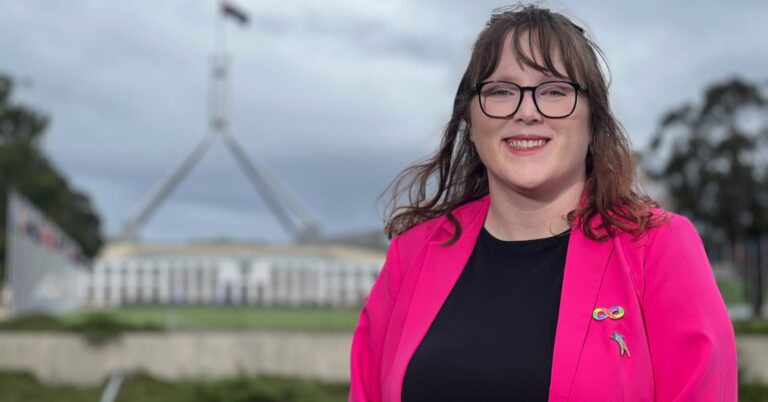 A young, smiling woman with fair skin and shoulder-length dark blond hair. She is wearing glasses, a black top and a pink blazer with an Autism Pride rainbow infinity pin on the lapel. She is standing in front of Parliament House in Canberra, a wide, low, white building with the Australian flag flying over it.