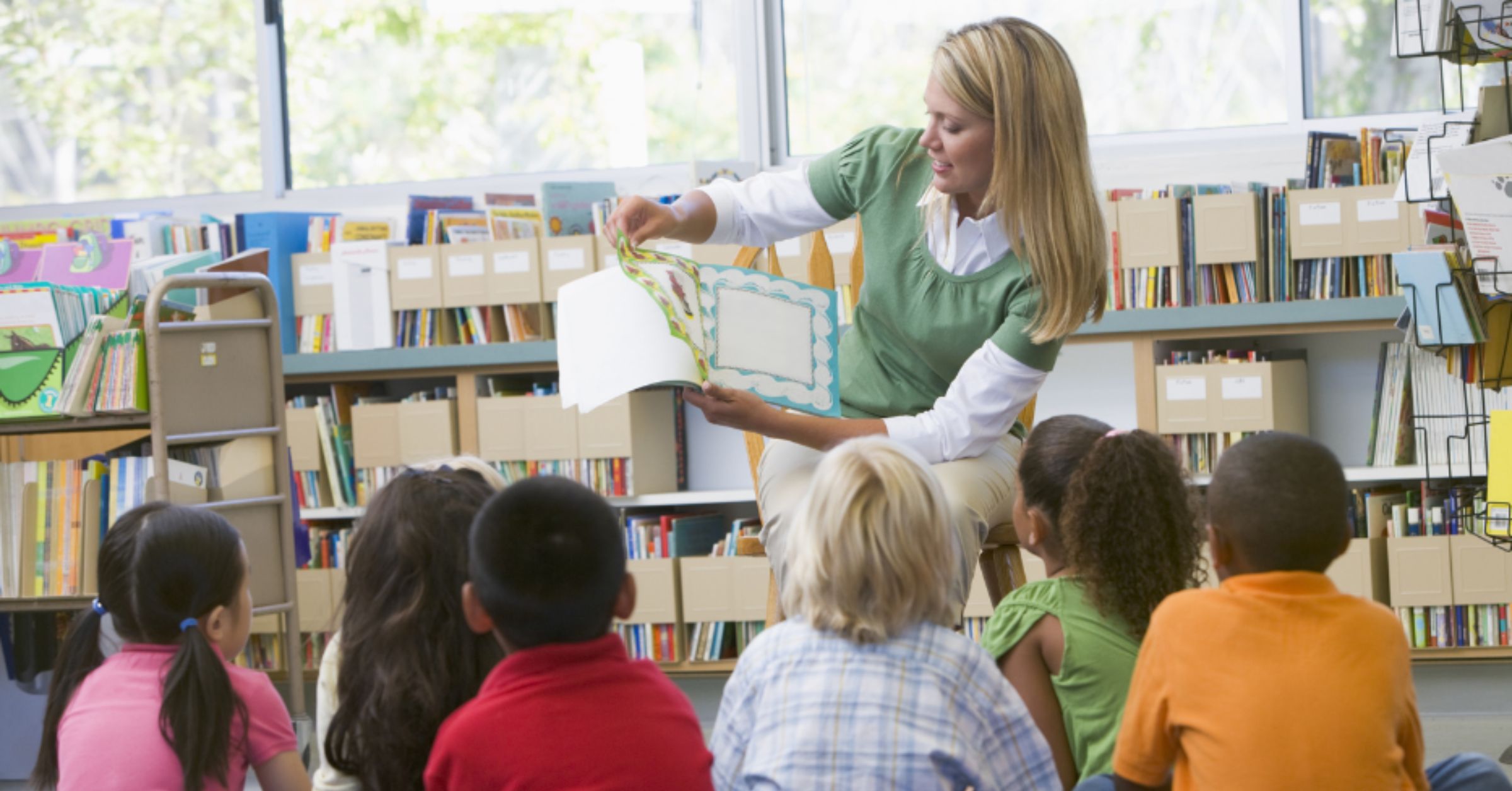 A photo of a teacher with long blonde hair reading from a picture book in front of a group of six young students. They are in a brightly lit classroom surrounded by bookshelves.