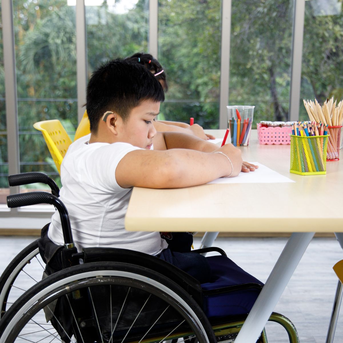 Young East Asian boy with short black hair wearing a white t-shirt and using a manual wheelchair and a hearing aid. He is at a table that looks a bit high for him, drawing with coloured pencils. There is another child at the table with him, mostly obscured by the boy due to the camera angle. Tthere are big windows behind them with a view of trees.