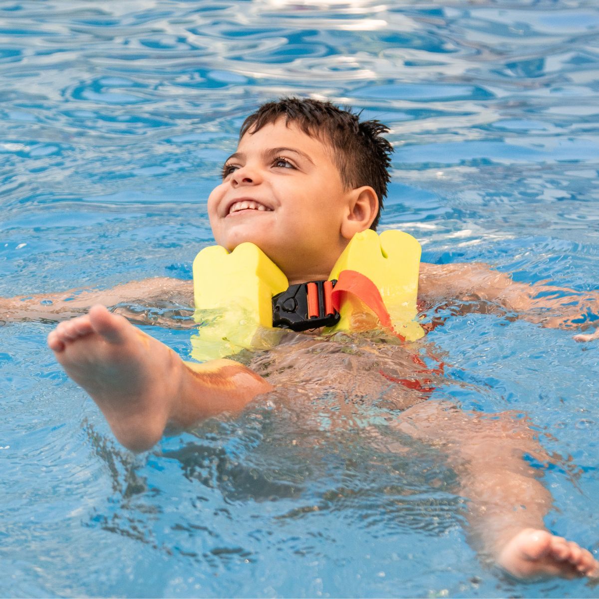 A young boy with olive skin and short brown hair wearing yellow floaties in a swimming pool. He is leaned back and kicking, smiling at something off to his right.