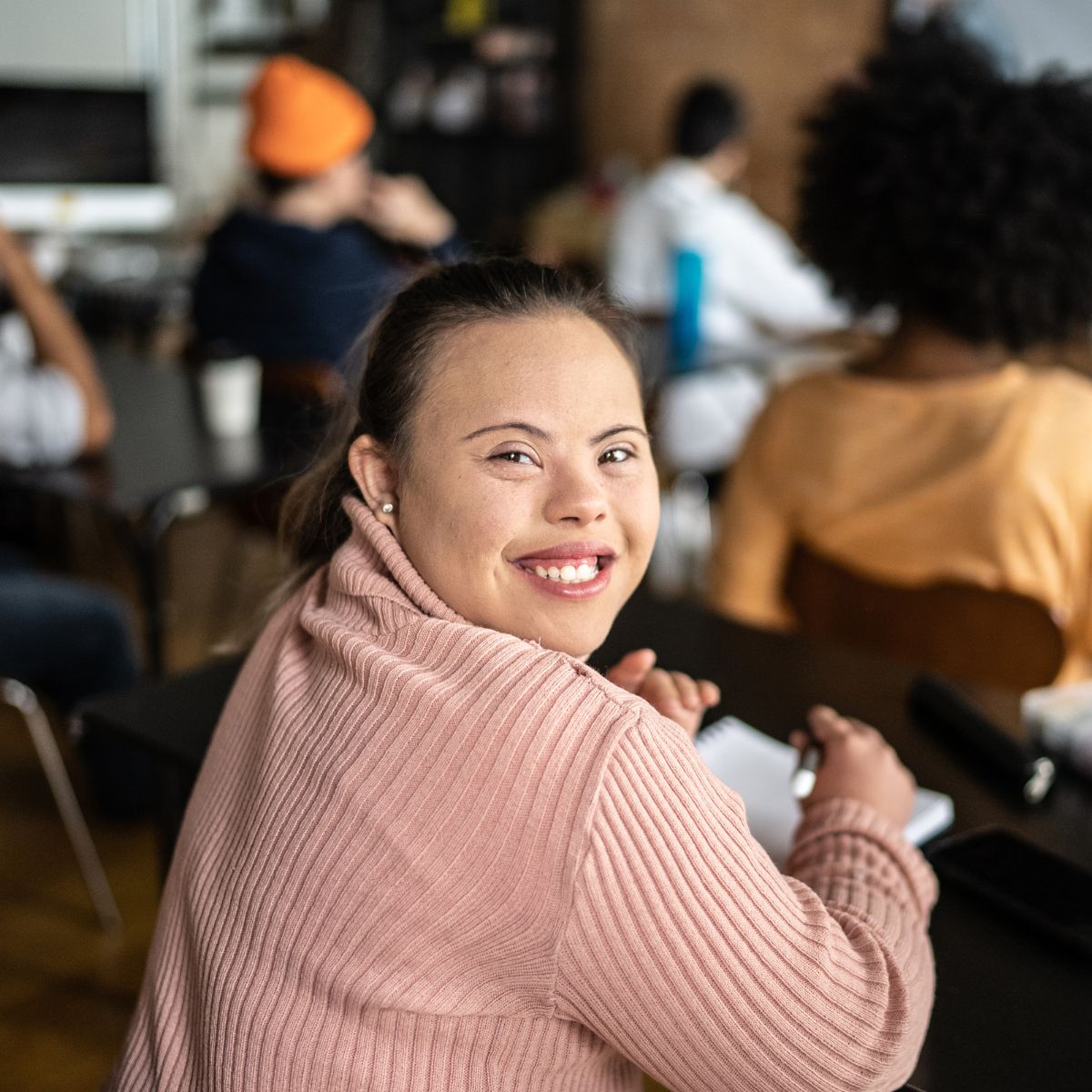 A young woman with fair skin and brown hair tied back in a ponytail wearing a knitted pink jumper. She has Down Syndrome. She is in a classroom of people studying and is twisted in her seat to grin at the camera behind her.