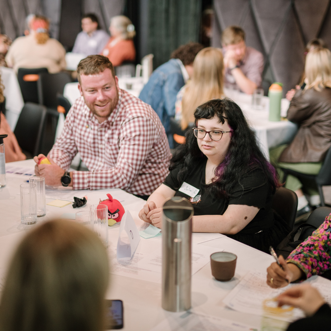 CYDA staff member Jase sits at a workshop table smiling and talking with a group of people