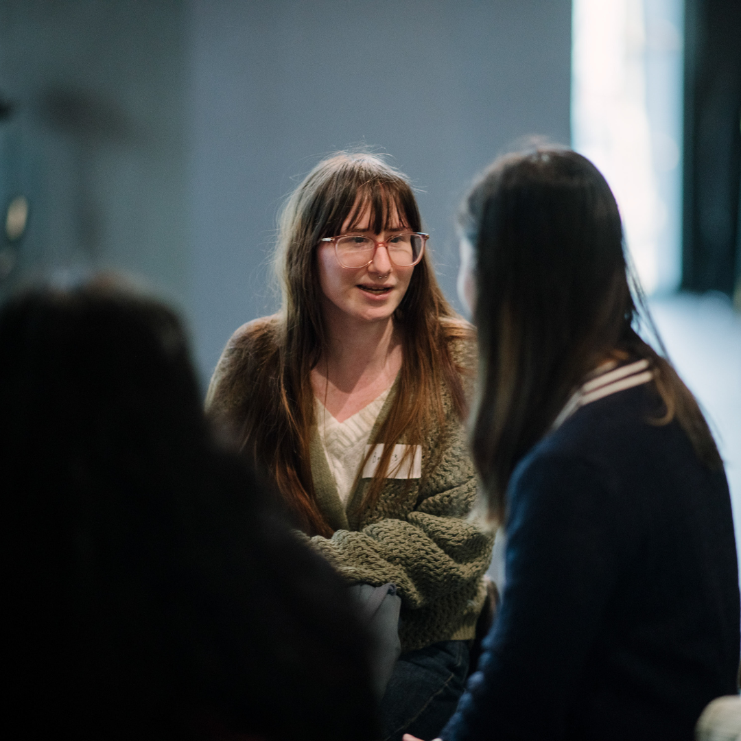 A young person with long brown hair and glasses talking to a group of people