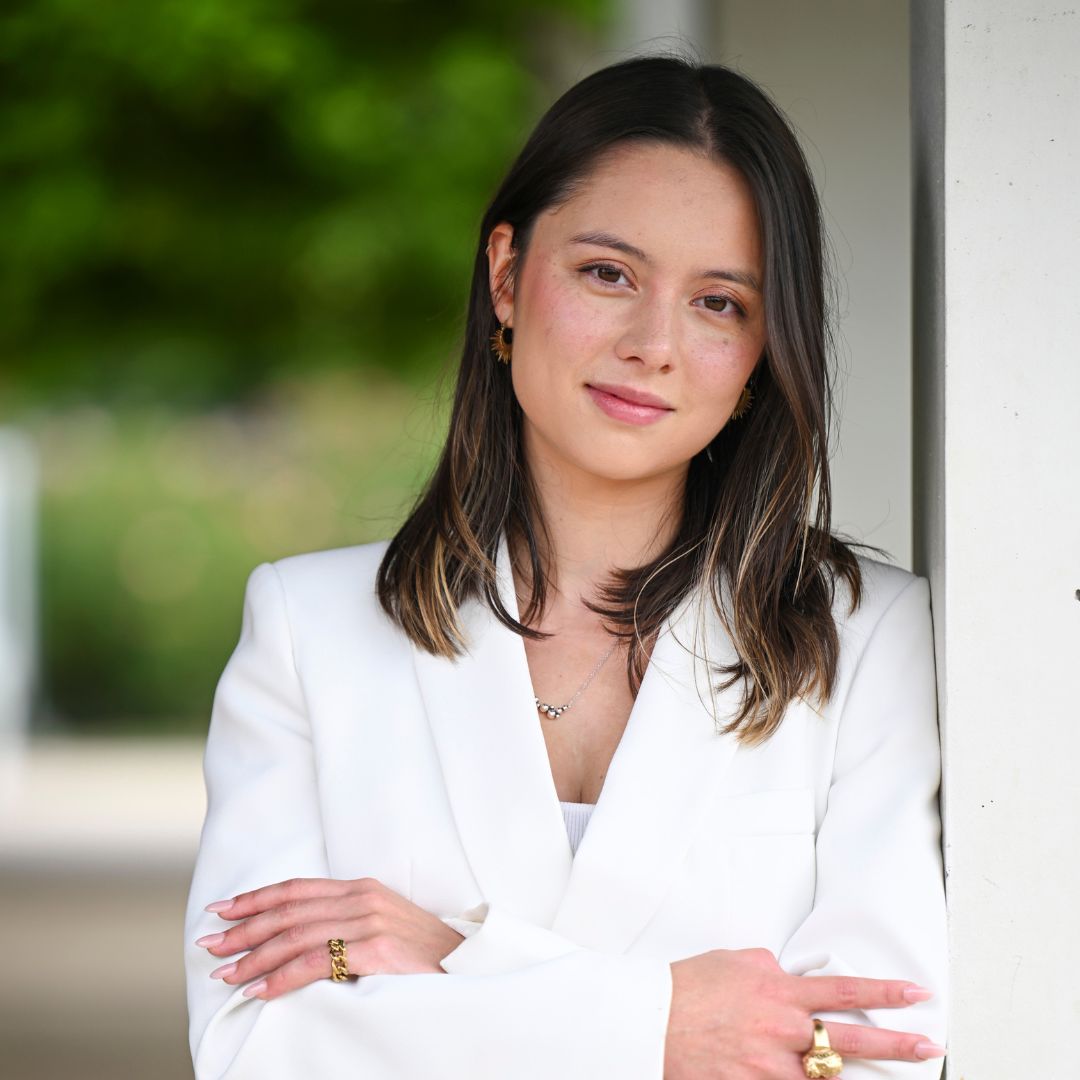 A photo of a young woman with long brown hair wearing a white suit jacket while leaning against a wall with her arms crossed. She has gold rings on both hands.