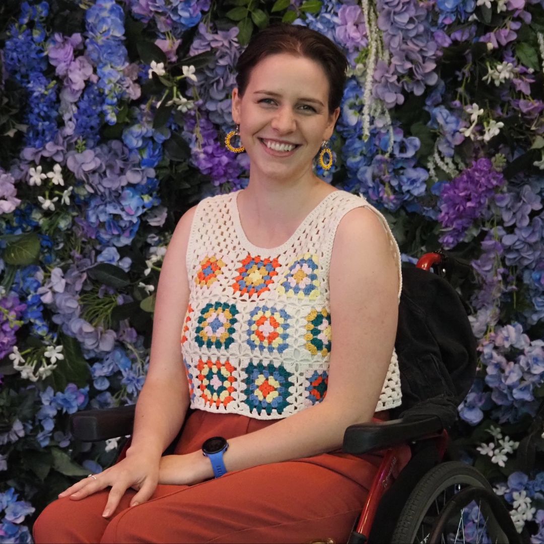 A photo of a smiling young person with short brown hair. They are wearing a white knitted top featuring colourful patterns and using a wheelchair. Purple flowers make up the background behind them.