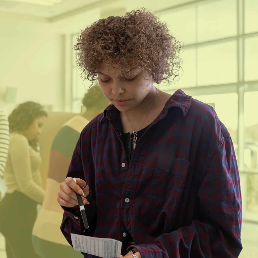 A photo of a young woman with curly brown hair wearing a red and purple button up shirt. She is at a polling station holding a pen as she looks down at a voting sheet.