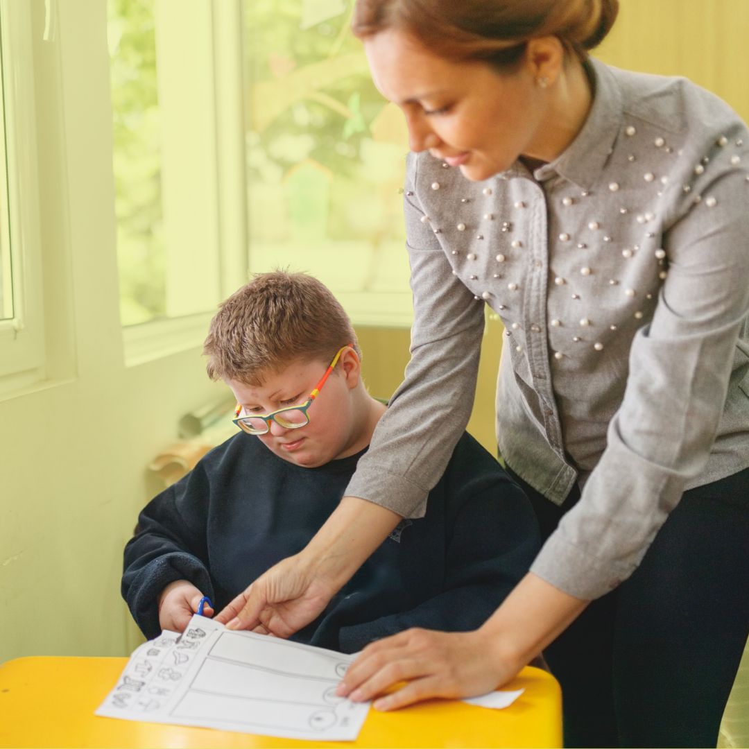 A photo of a boy with short blond hair wearing glasses and a black sweater. He is using a pair of blue scissors to cut a worksheet on the desk in front of him. His teacher, a woman in a grey top and black pants, is standing beside him while holding the paper as he cuts.