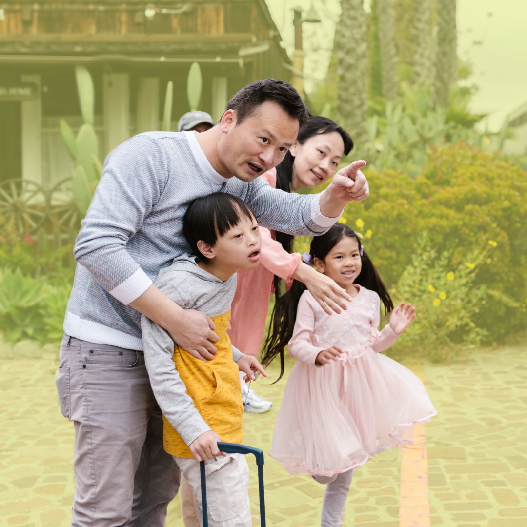 A photo of a family of four. A boy and a girl stand with their father and mother in front of green foliage. The father is pointing ahead of the group.