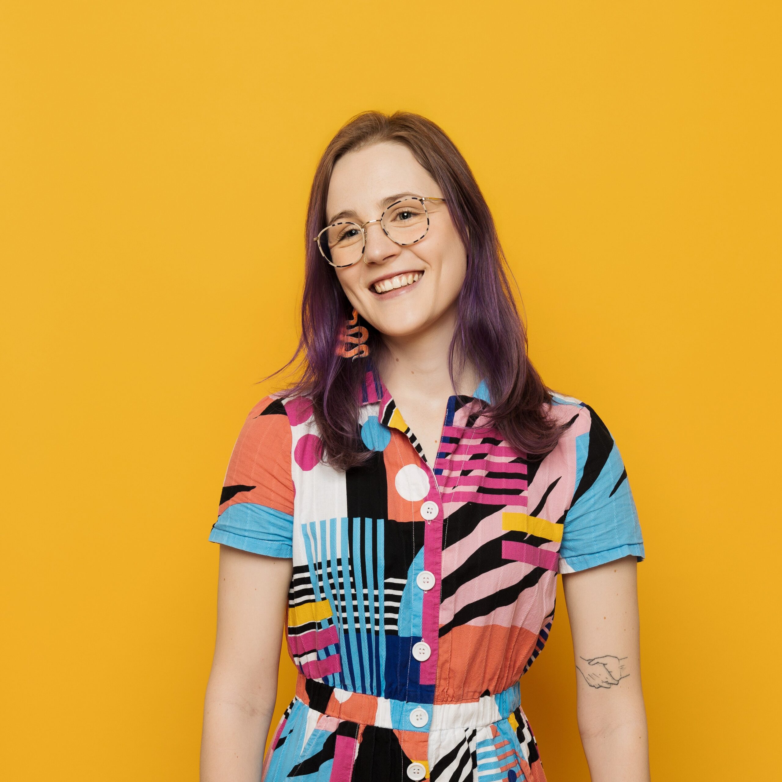 A photo of a young person with long brown hair wearing colourful clothes standing against a yellow wall.