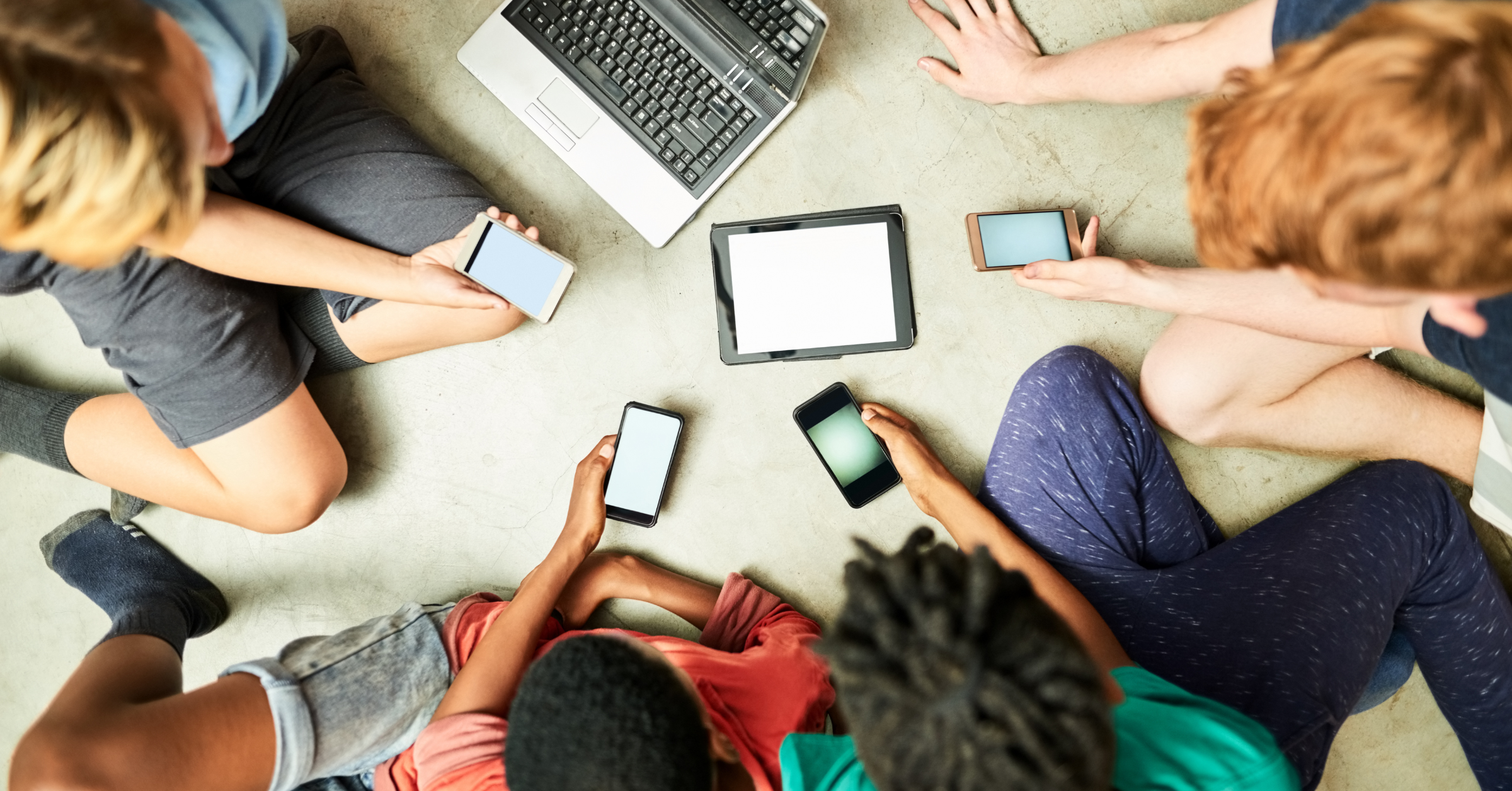 A group of teenagers sitting on the floor. Between them lie several smartphones, a tablet, and an open laptop.