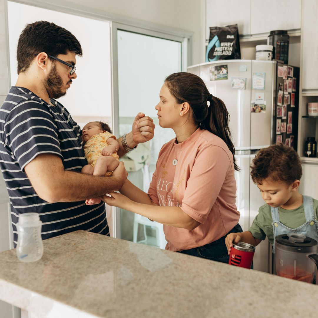 A photo of a family in a kitchen. A woman with long brown hair is handing a baby to a man in a striped t-shirt. A boy the right is looking at a cup.