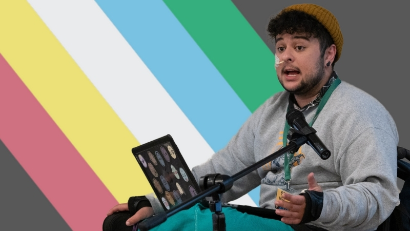 Photograph of a young man with olive skin and dark curly hair and a short beard sitting with a laptop on his lap and speaking into a microphone. He is wearing a mustard beanie, a green lanyard and has a feeding tube in his nose. The photograph is superimposed over the disability pride flag.