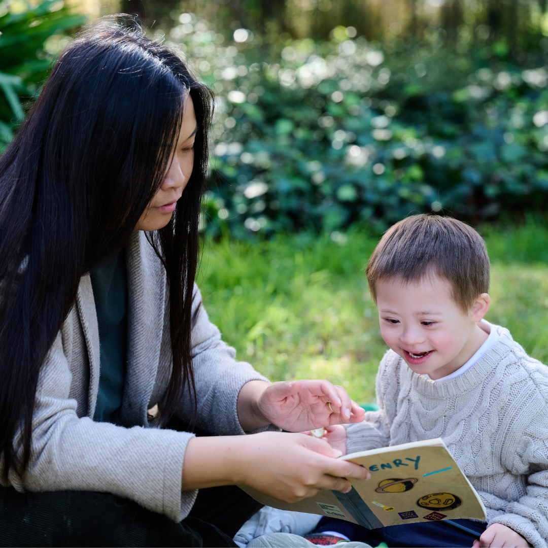 A mum and a young child with down syndrome reading a book together in a park.